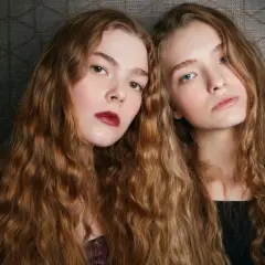Dwie dziewczyny z długimi, falowanymi włosami. Czy farbowanie niszczy włosy? Ich fryzury wyglądają na zdrowe i lśniące.