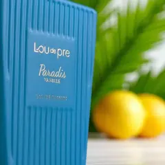 Niebieska butelka perfum Lou de Pre Paradis Vanille na tle liści i cytryn.