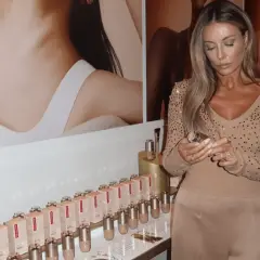 Kobieta z długimi blond włosami trzyma buteleczkę Clarins Double Serum Foundation. Na półce stoją dziesiątki opakowań tego podkładu.