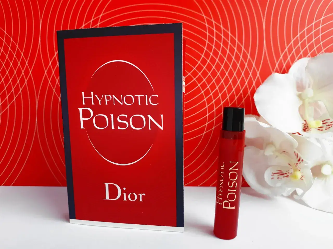 Czerwone opakowanie i flakonik perfum Christian Dior Hypnotic Poison na tle czerwonej, falistej tapety i białych kwiatów.